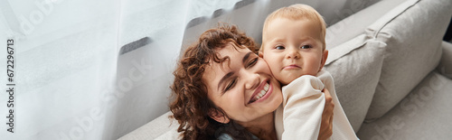 happy mother with closed eyes holding adorable toddler child on couch in living room, banner