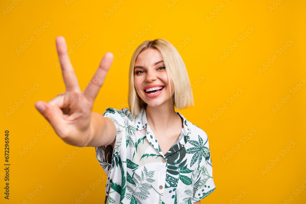 Photo portrait of pretty young girl show v-sign toothy smile dressed stylish leaves print outfit isolated on yellow color background