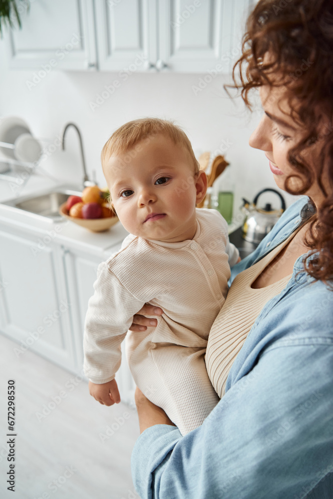 happy woman holding cute little daughter in romper while standing in kitchen at home, loving mother happy woman holding cute little daughter in romper while standing in kitchen at home, loving mother