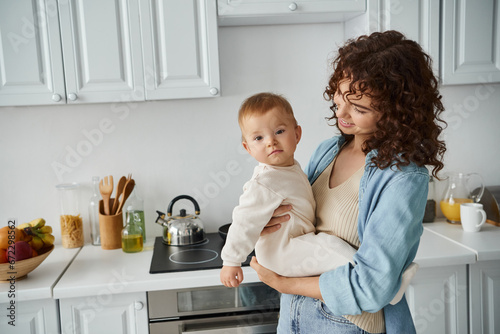 smiling curly woman holding baby girl in romper looking at camera in cozy kitchen, happy family