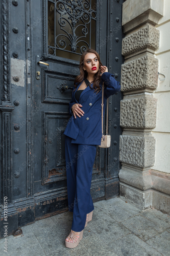 Trendy beautiful stylish chic urban woman with red lips in fashion blue suit with bag and shoes stands and poses near a vintage black door on the street. Pretty elegant lady Trendy beautiful stylish chic urban woman with red lips in fashion blue suit with bag and shoes stands and poses near a vintage black door on the street. Pretty elegant lady
