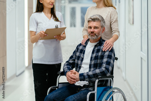 Grey haired patient in wheelchair in hospital. Charming wife pushing wheelchair with husband consulting doctor at rehab center. © Iryna