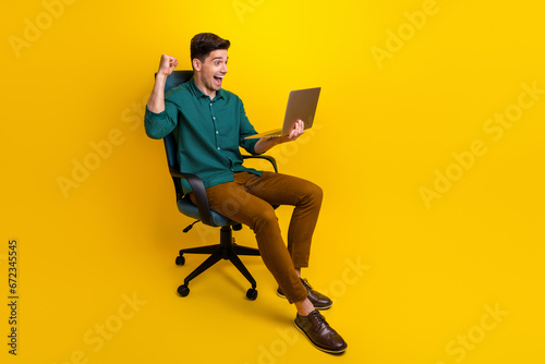 Photo of overjoyed glad man sitting armchair watching video showing hand fist rejoice sale empty space isolated on yellow color background