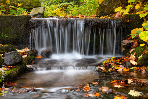 Small cascade in Sauerland Germany from frog perspective with long time exposure. Brook falling down in autumn scenery with colorful leaves of oak trees. Idyllic October atmosphere in park in Iserlohn