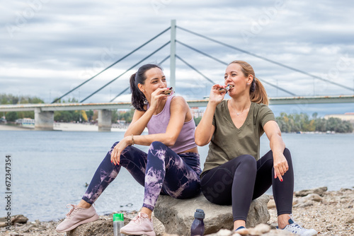 Two female friends pursuing their activity outdoors. Resting by the river bank after jogging, and refreshing with a healthy snack or a cereal bar.