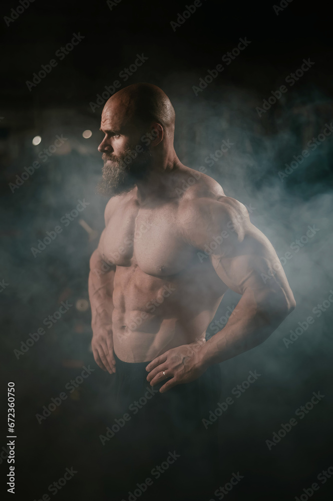 A muscular bald man poses shirtless in the dark amid smoke. A bodybuilder shows off his form in the gym.