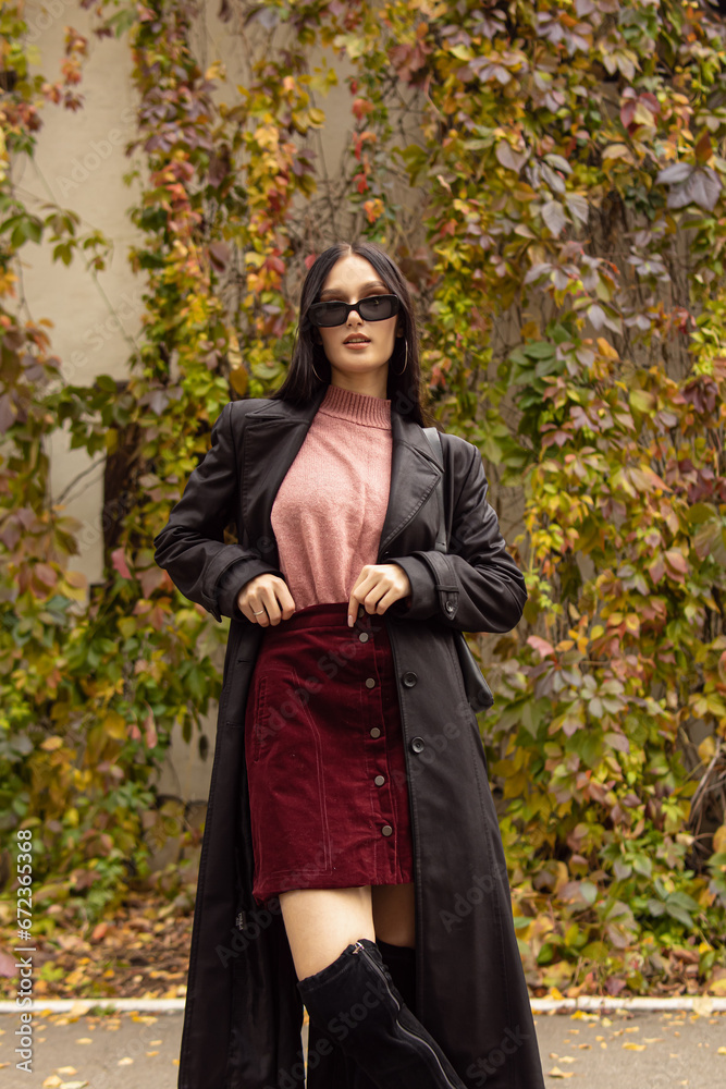 Young woman in black sunglasses, leather raincoat, pink turtleneck and red skirt. Natural. Attractive woman in park. Youth fashion Young woman in black sunglasses, leather raincoat, pink turtleneck and red skirt. Natural. Attractive woman in park. Youth fashion