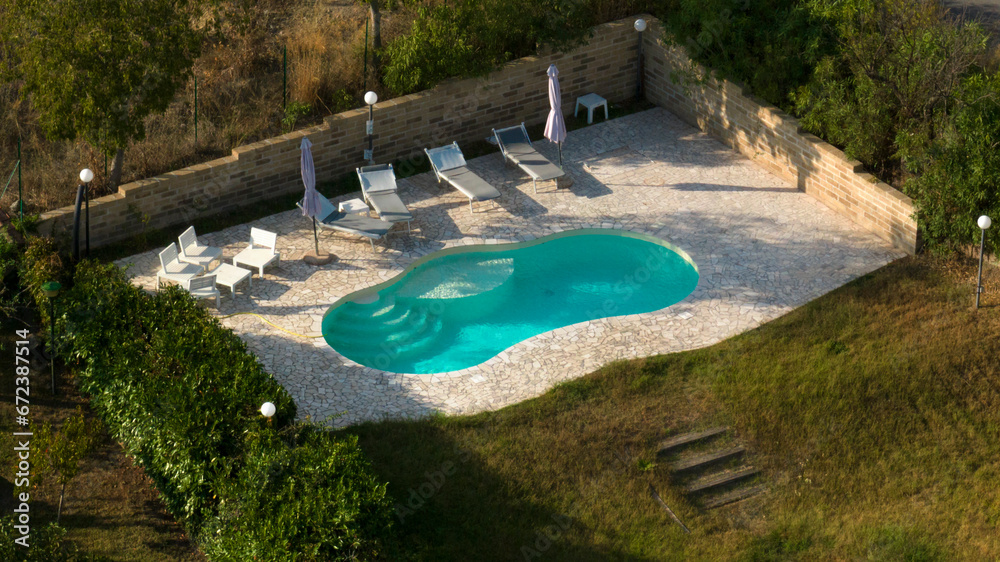 Aerial view of a swimming pool with ladder, belonging to a large villa. The pool is empty and no one is swimming. Around the water there is a stone floor.