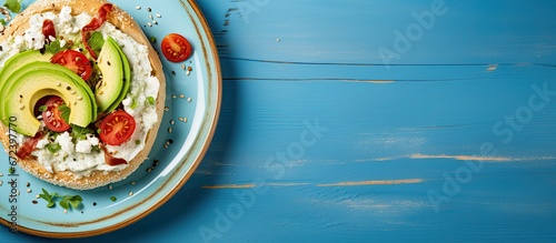 Ceramic plate with a flat lay presentation on a blue texture background featuring a whole wheat bagel sandwich filled with vegetarian ingredients like chopped avocado cream cheese sun dried 
