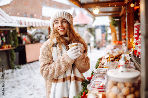 Happy woman drinks a hot drink from a glass and walks through the Christmas market on a sunny winter day. Spending winter holidays. Holiday concept.