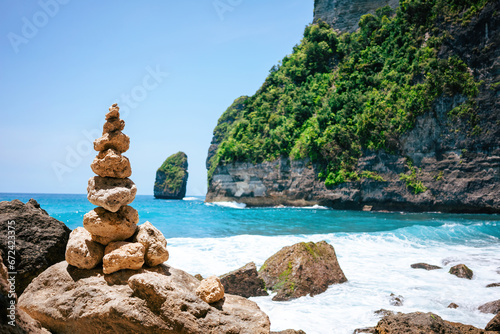 Zen rock tower stands at the forefront of a breathtaking tropical coastline. Behind, emerald waves crash against rugged cliffs adorned with green foliage.