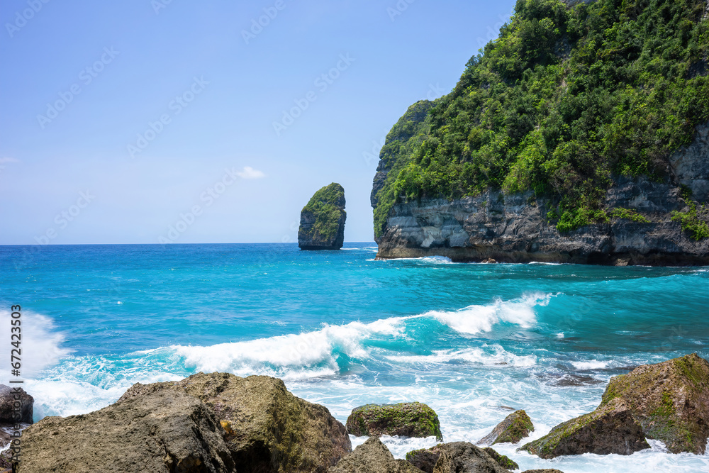 Turquoise waves crash against the shoreline, with a lush green cliff and unique rock formation looming in the distance. Clear skies and vibrant waters create a picturesque paradise