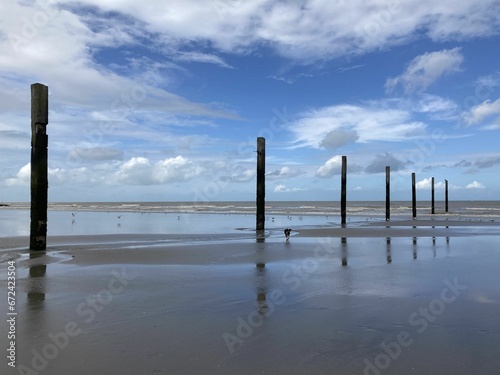 Beach with wooden posts extending out into the crystal blue waters against a cloudy blue sky