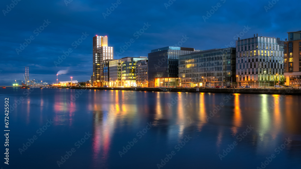 Modern offices and apartment buildings on Sir John Rogerson Quay and Poolbeg Powerstation chimneys, blurred Liffey River at dusk, Dublin, Ireland