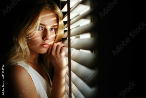 jeune fille blonde regardant à travers un store à lamelles dans la pénombre de son appartement