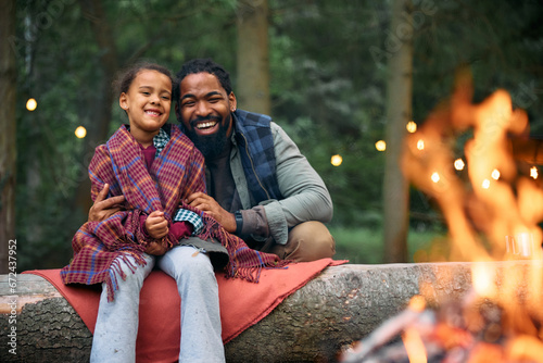 Happy black father and daughter enjoy by bonfire while camping in woods. photo