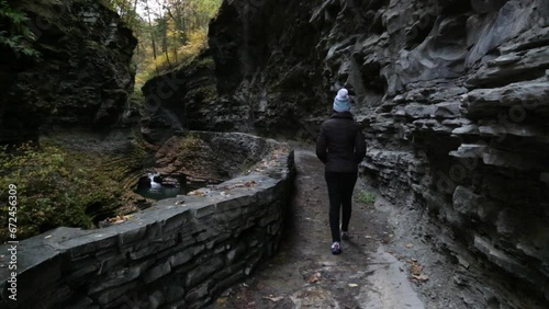 woman walking along path in watkins glen state park (waterfall trails with autumn foliage, leaves turning colors in fall) dressed in jacket, hat, wet ground, stone walkway inside gorge, finger lakes photo