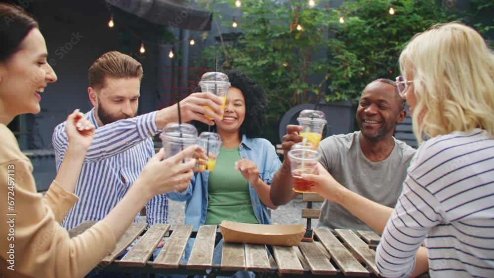 Good-looking mixed-raced friends celebrating meeting on bar terrace. Cheerful people clinking cocktails and laughing loudly while sitting at table. Actively speaking with each other at cafe outside.
