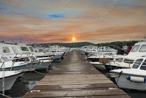 View along a wooden jetty in a harbor with docked ships in front of the setting sun © Aquarius