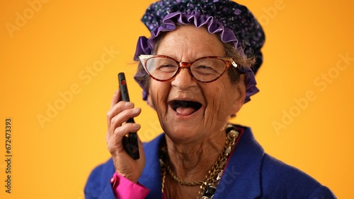 Fotografie, Obraz Closeup of funny crazy happy elderly old toothless woman talking on mobile cell phone wearing glasses isolated on solid yellow background studio portrait