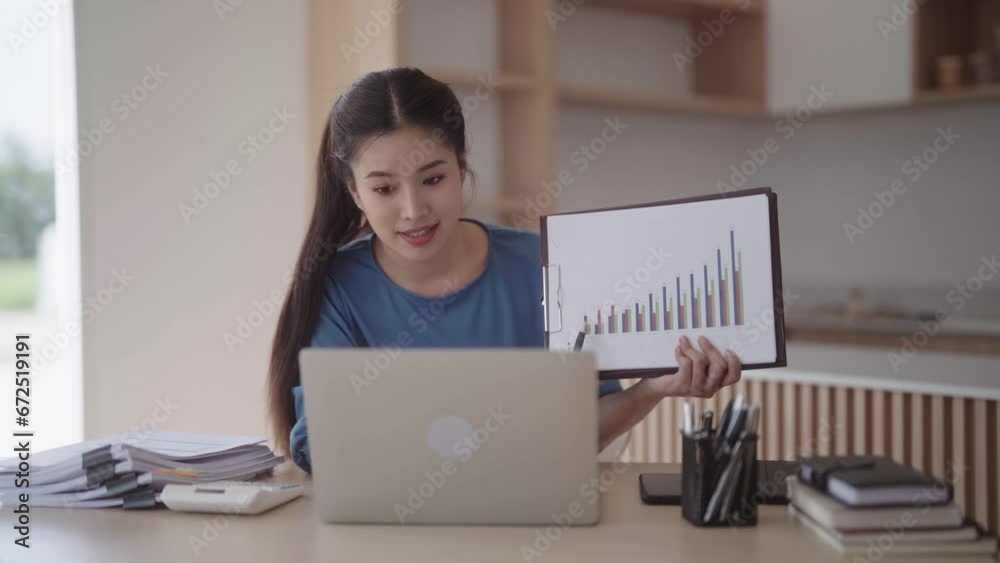 Asian woman presenting and explaining profit and loss graphs through the internet using her laptop computer. She is at her dining table in a modern minimalist-style kitchen at her home.