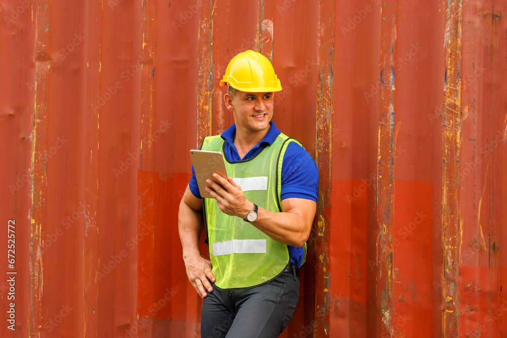 Caucasian man wearing yellow safety helmet Wear a reflective safety suit, hold a tablet, and stand smiling against a container. In the container storage yard Caucasian man wearing yellow safety helmet Wear a reflective safety suit, hold a tablet, and stand smiling against a container. In the container storage yard