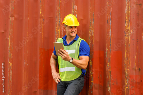 Caucasian man wearing yellow safety helmet Wear a reflective safety suit, hold a tablet, and stand smiling against a container. In the container storage yard