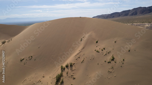 A large sand dune in the middle of the steppe. Drone view of a huge pile of sand. Tourists are walking  enjoying the view. A river runs in the distance and grass grows. The sky with white clouds