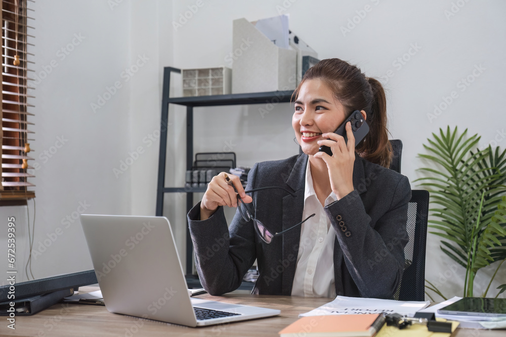 Businesswoman talking on business phone and using laptop to do finance, mathematics on wooden table in office and business background, tax, accounting, statistics and analytical research concepts. Businesswoman talking on business phone and using laptop to do finance, mathematics on wooden table in office and business background, tax, accounting, statistics and analytical research concepts.