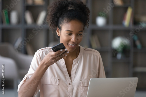 Happy young African American woman recording and sending audio message on mobile phone, using voice recognition app. Gen Z student girl giving command to virtual assistant on mobile phone at laptop