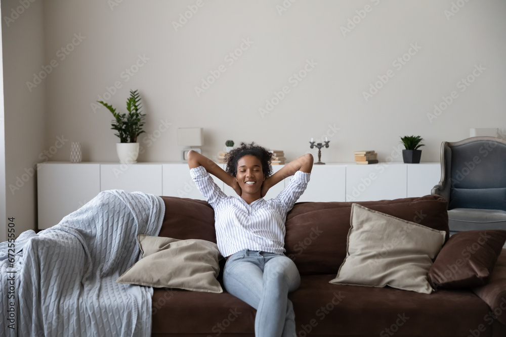 Happy young African American woman relaxing on sofa at home, resting on couch, breathing cool fresh conditioning air, enjoying leisure time in ventilated living room. Comfortable house concept
