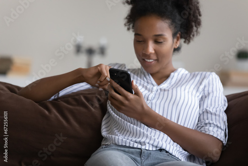 Happy relaxed young Black girl, woman resting on couch, enjoying leisure, using app on smartphone, reading book online, making phone call, watching video, browsing internet