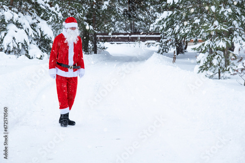 Santa Claus is stands on a snowy fairy-tale road outdoor in winter with pine trees. Celebrating Christmas and New Year.