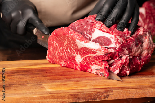 The butcher wearing black gloves cuts the meat with knife on wooden cutting board.Close-up piece of raw meat with a chef's knife on a dark background. photo
