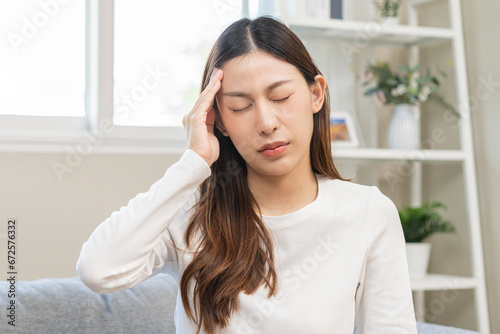 Dizzy asian young woman, girl headache or migraine pain suffering from vertigo while sitting on couch in living room at home, holding head with hand, health problem of brain or inner ear not balance.