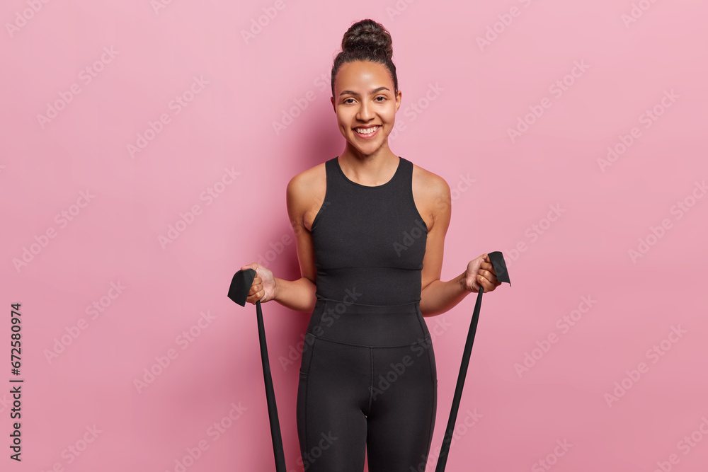 Happy sportswoman doing exercise with resistance band working out in gym smiles happily dressed in black tracksuit isolated over pink background being in good physical shape. Be healthy and strong