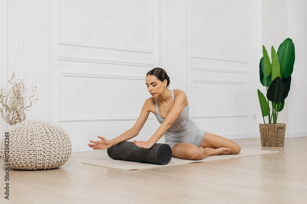Attractive slim girl using a roller to relax her body muscles after a workout. A young fit woman in light sportswear posing against a light background. Attractive slim girl using a roller to relax her body muscles after a workout. A young fit woman in light sportswear posing against a light background.