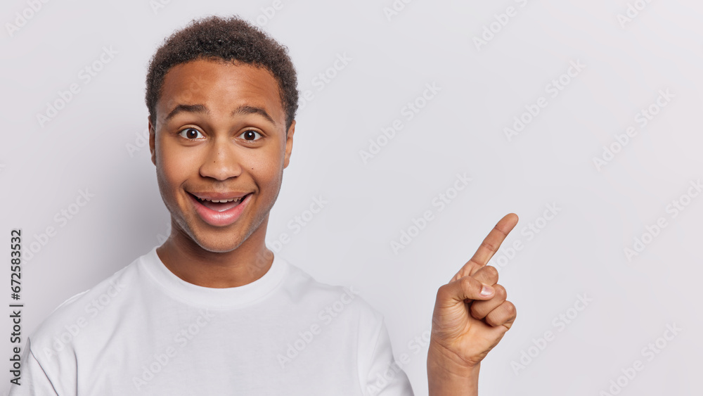 Handsome curly haired African man dressed in casual clothing points index finger at blank space for your promotional content keeps mouth opened isolated over white background. Hey look at this Handsome curly haired African man dressed in casual clothing points index finger at blank space for your promotional content keeps mouth opened isolated over white background. Hey look at this