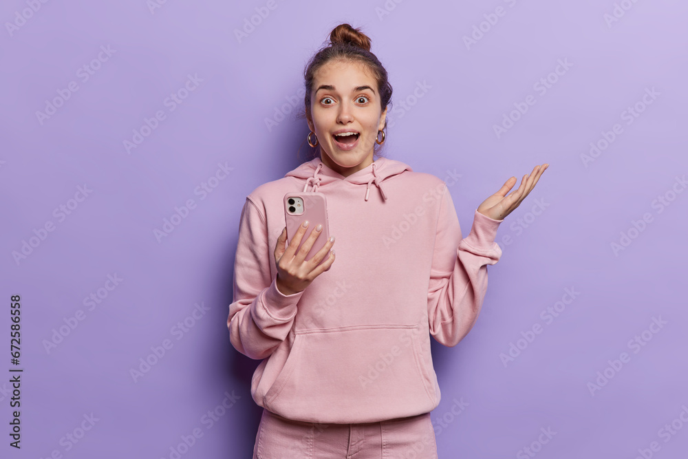 Young suprised woman with hair bun reacts surprisingly to something holds breath reacts to something amazing uses mobile phone dressed in casual hoodie isolated over purple studio background