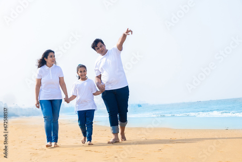 Happy indian Family of three enjoying summer vacation. father and mother holding hands of their daughter and pointing on beach.