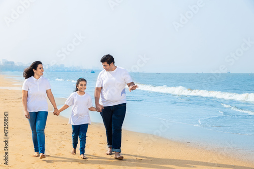 Happy indian Family of three enjoying summer vacation. father and mother holding hands of their daughter on beach.