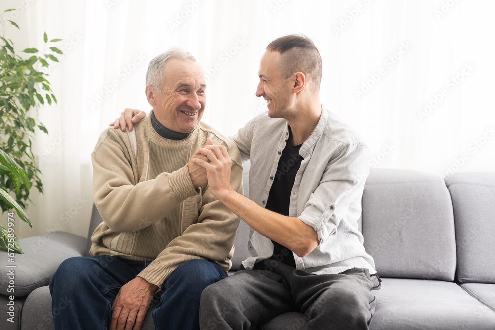 Happy two generations male family old senior mature father and smiling young adult grown son enjoying talking chatting bonding relaxing having friendly positive conversation sit on sofa at home. Happy two generations male family old senior mature father and smiling young adult grown son enjoying talking chatting bonding relaxing having friendly positive conversation sit on sofa at home.