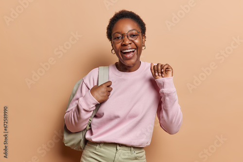 People positive emotions concept. Indoor waist up of young happy smiling broadly African american female student standing in centre on beige background wearing casual clothes and bag on shoulder photo