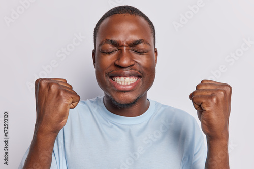 Happy thrilled dark skinned man with small beard clenching fists celebrating success enjoying victory keeps eyes closed shows even teeth isolated over white background. Empowered triumph concept