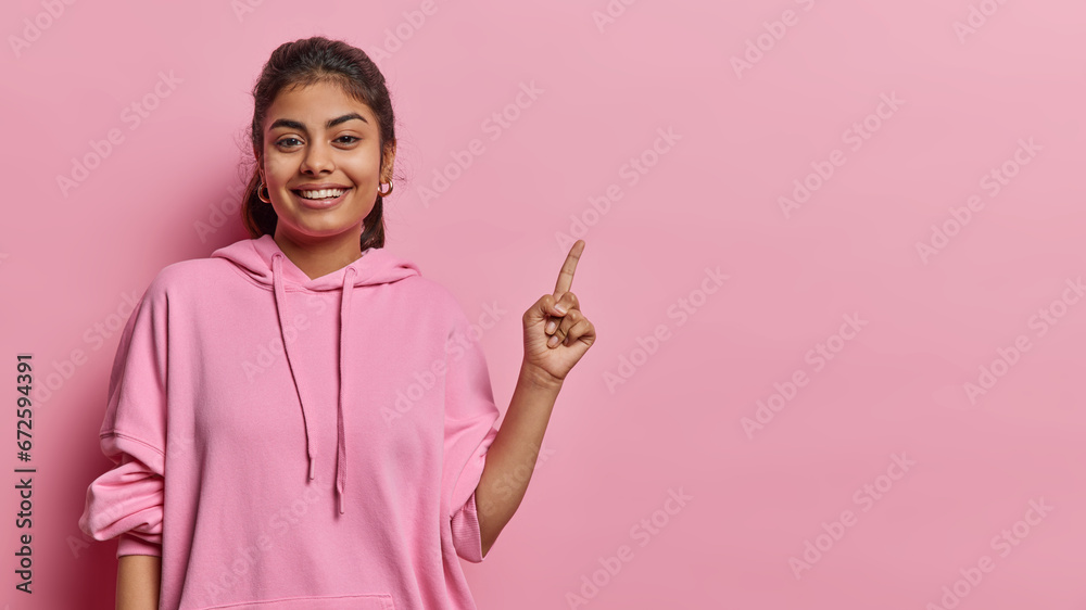 Horizontal shot of pleased Iranian woman with dark hair smiles gladfully dressed in casual sweatshirt points index finger on copy space isolated over pink background. Place your advertisement here Horizontal shot of pleased Iranian woman with dark hair smiles gladfully dressed in casual sweatshirt points index finger on copy space isolated over pink background. Place your advertisement here