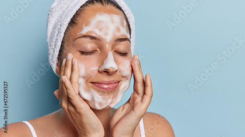 Proper hygiene and pampering concept. Headshot of young woman has facial cleansing routine enjoying moment of relaxation during her skincare has white soft towel on head isolated over blue background