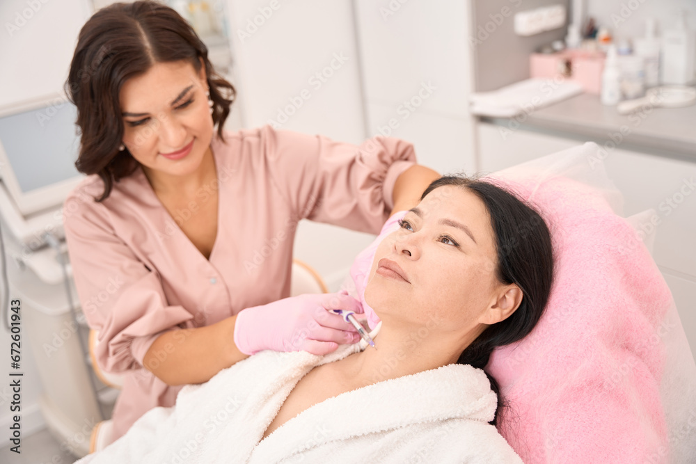 Female cosmetologist gives beauty injections to the neck area of an Asian client Female cosmetologist gives beauty injections to the neck area of an Asian client