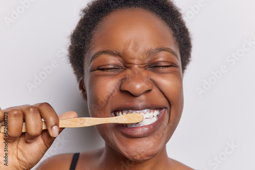 Teeth hygiene concept. Close up shot of dark skinned woman with curly hair cleans teeth holds wooden toothbrush has eyes closed isolated over white background. Morning procedure and perfect smile photo