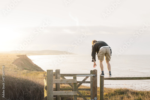 A man climbing a fence on a clifftop with a sunning sea view photo