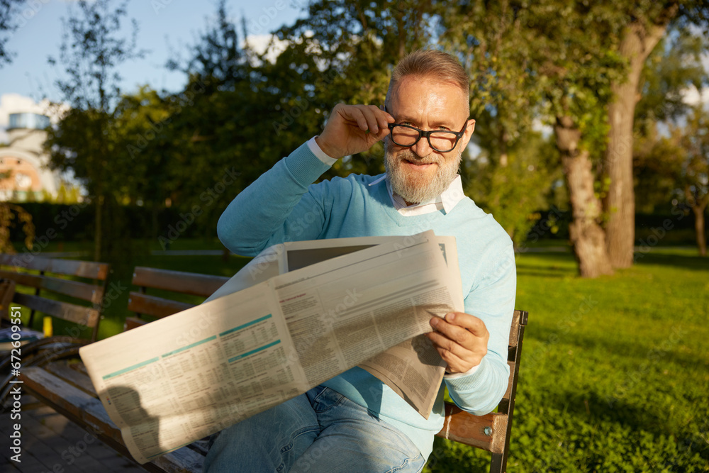 Fototapeta premium Confident bearded mature man reading daily newspaper on park bench